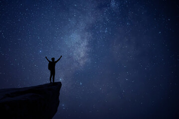 Silhouette of young man standing alone on top of mountain and raise both arms enjoying nature on beautiful night sky, star, milky way background. Demonstrates hope and freedom.