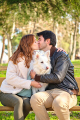 Young caucasian couple kissing with their dog sitting on a bench in a park. Family.