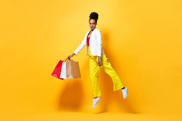Chic stylish African American woman jumping with shopping bags in yellow color isolated background...