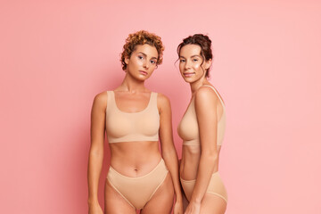 Two beautiful girls in beige lingerie stand against pink wall, one girl front view, other girl head to shoulder , friendship concept, copy space - obrazy, fototapety, plakaty