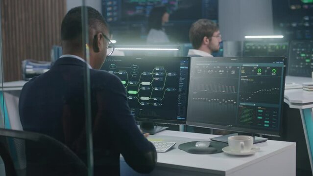 African American big data scientist works at computer in monitoring room. Multiracial IT engineers analyze charts at background. Multiple big screens on the wall with displayed blockchain network map.