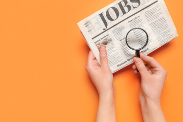 Female hands with mini magnifier and newspaper with headline JOBS on orange background. Search...