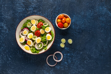 Tasty salad with quail eggs, tomatoes, onion and lettuce on dark blue background