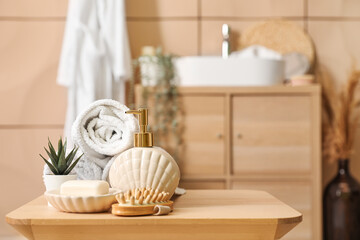 Soap dispenser, bar, massage brush, rolled towels and houseplant on wooden table in bathroom,...