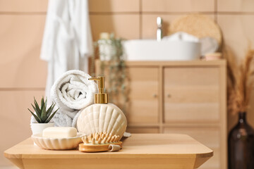 Soap dispenser, bar, massage brush, rolled towels and houseplant on wooden table in bathroom, closeup