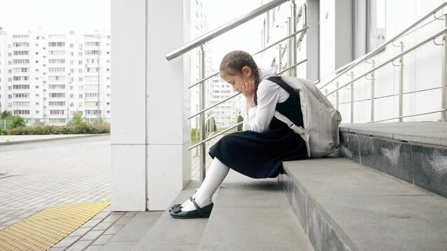 Brown-haired girl sits on empty entrance staircase without desire to attend lessons at school. Junior schoolgirl sits with sad expression