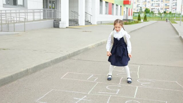 Junior schoolgirl playing hopscotch in empty school yard. Brown-haired girl in uniform with backpack enjoys playing alone at school, slow motion