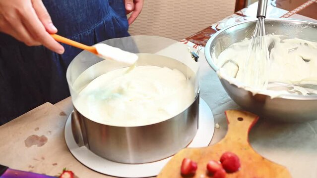 Close-up of a housewife, chef confectioner spreading whipped cream, smoothing the top layer of the mousse cake inside a removable metal mold, preparing delicious light French summer dessert Fraisier