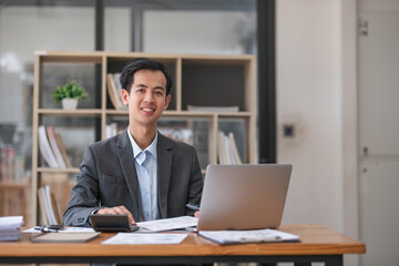 A handsome and smart millennial Asian businessman or male boss in a formal business suit focuses on his work on laptop computer in his office.