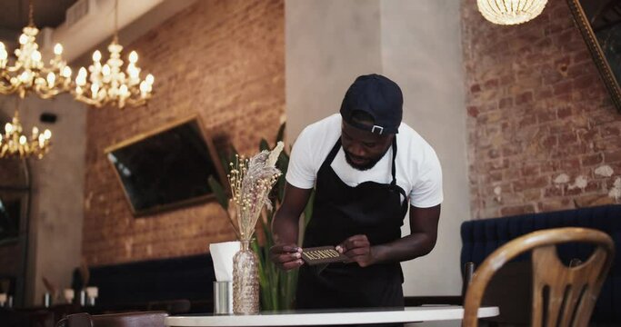 A Black person in a black apron and white T-shirt places a Reserved sign on a table in a restaurant. Table reservation in a restaurant