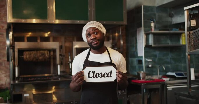 A Black person in a doner market is holding a Close sign and is sad because the doner market is closing
