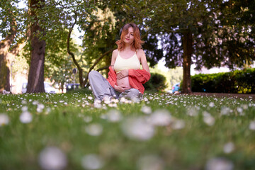 Young pregnant woman applying headphones on belly to listen to music in shady park on the summer day.regnancy and maternity concept.Pregnancy women is relaxing with music and headphone in outdoor.