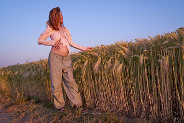  A pregnant woman is holding her stomach in the middle of a wheat field. A girl is expecting a baby at sunset in summer. Fatherhood and motherhood among young people
