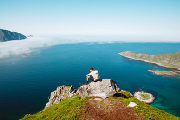 Young athletic man sit on rock on top of mountain or hiking trail overlooking ocean. Man with backpack and cap after long summit rest on top enjoy the view 