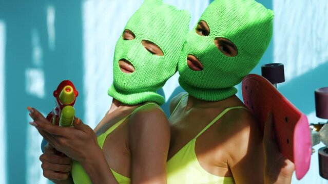 Two beautiful sexy women in green underwear. Models wearing bandit balaclava mask. Hot seductive female in nice lingerie near blue wall. Crime and violence. With penny skateboard  and water gun