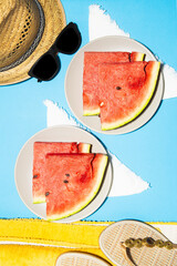 Top vertical view of two plates with slices of watermelon on blue background, straw hat, sandals and beach towel
