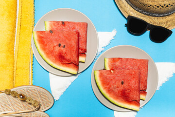 Top view of two plates with slices of watermelon on blue background, straw hat, sandals and beach towel