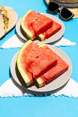 Front vertical view of two plates with slices of watermelon on blue background, straw hat. sandals and sunglasses