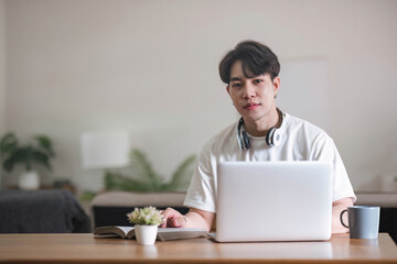 A focused young Asian male college student or freelancer using his digital tablet at a table in his living room.