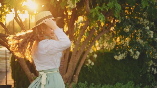 Cheerful happy joyful young woman red long loose hair fly wind throws. cute Girl spinning dancing hands raised to magic sun light flare sunset. Beauty Smiling face. summer nature reen trees. boat hat 