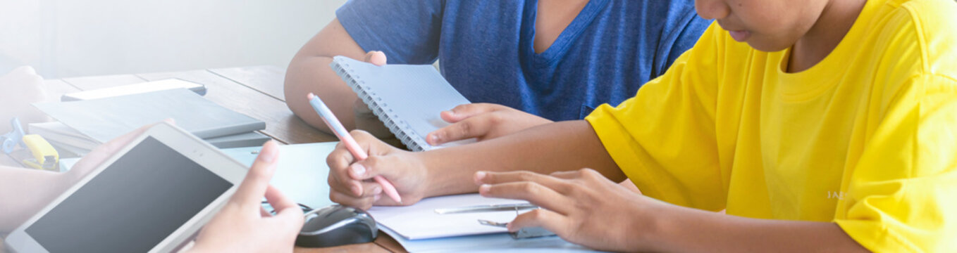 Closeup image of hands of young asian boys who are using tablet, pens, mobile phone, headphone and other learning tools on table, cropped shot, soft and selective focus.