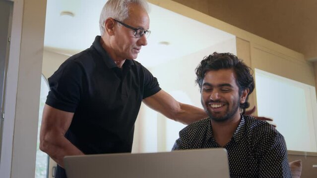 An old Indian boss is examining his employee or subordinate's work on a laptop in an office space and appreciates him with a pat on his back, making the employee happy who is smiling as his boss leave