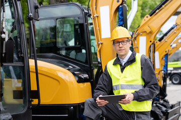 Engineer in a helmet with a digital tablet stands next to construction excavators.