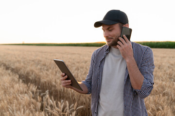Bearded farmer with mobile phone and digital tablet in agricultural field at sunset.