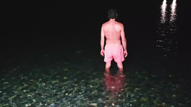A man in swimming shorts dives into the night sea. Multicolored sea stones under clear water on a pebble beach at night with artificial light
