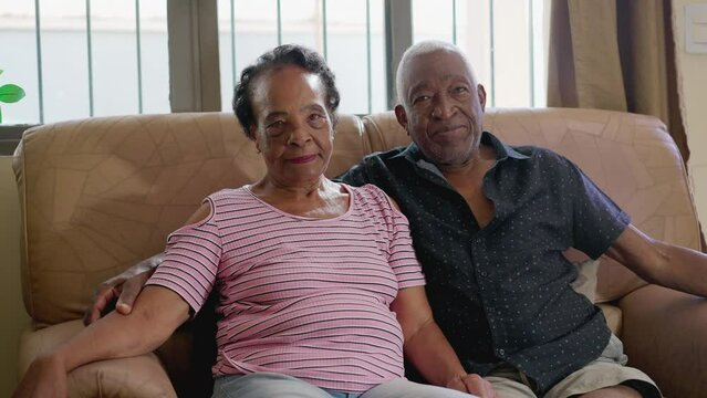 Portrait of a senior couple seated on couch looking at camera with neutral expression. A retired black older husband and wife together