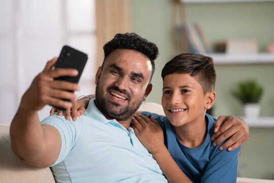 Happy smiling indian father with son taking selfie on mobile phone on sofa at home - concept of social media sharing, cyberspace and family time.