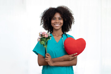 Young woman in blue scrubs uniform and stethoscope and standing holding heart shape with tulips...