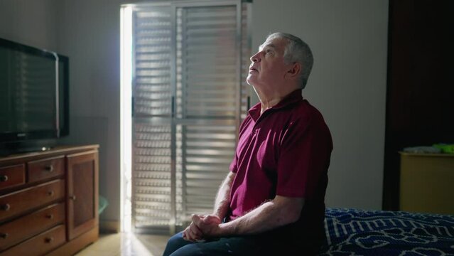 Religious Catholic senior man doing the sign of the cross in PRAYER sitting by bedside at home. Spiritual person having GRATITUDE and FAITH