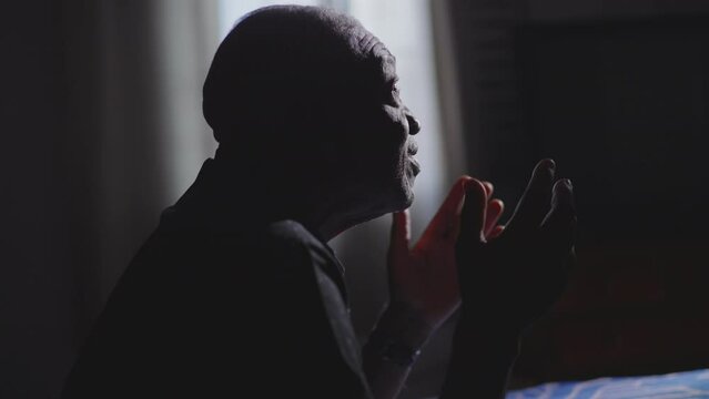 Religious African American Senior man kneeling by bedside PRAYING to GOD asking for help and GUIDANCE. Candid person looking up at sky with HOPE and FAITH in bedroom