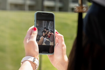 Photo on screen and hands of person wearing graduation cap holding a phone to take a group selfie photo with fellow graduates