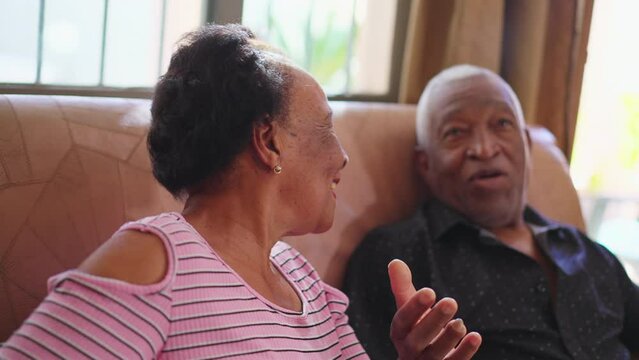 Senior African American couple sitting on couch interacting. A Brazilian older woman turning head to camera smiling feeling happy, old age retirement relationship