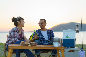 Asian couple drinking beer from bottle in their camping area with lake in the background during sunset...