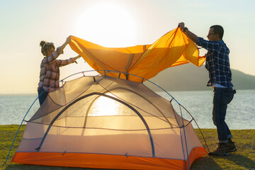 Asian couple preparing a tent to camping in the lawn with the lake in the background during sunset..