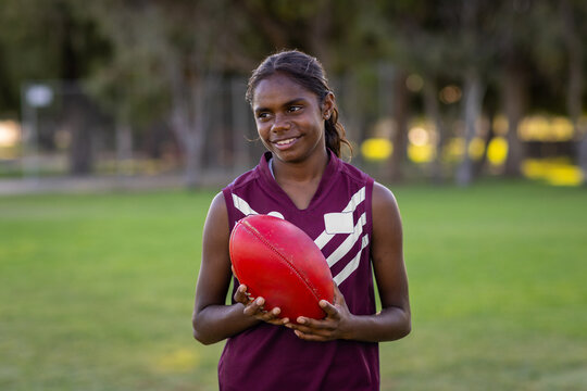aboriginal girl holding red leather football on grassed playing field