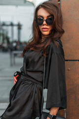 Fashionable young elegant pretty woman model with fashion sunglasses in elegant black suit with handbag stand near a wall in the city