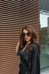 Beautiful trendy chic girl with cool sunglasses in a fashion black suit near a metal rust wall walks on the street