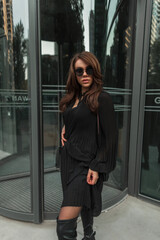 Fashionable beautiful glamor stylish street woman with cool sunglasses in a trendy fashion black dress stands near a modern building in the city. Pretty lady