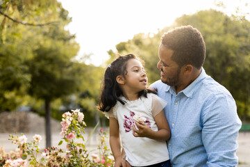 A dark-skinned father poses with his daughter near a park garden at dusk. The African adult looks happily at the little girl who has a flower in her hand. Concept of African-American parents.