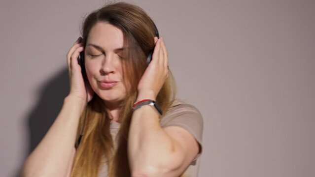 young woman with long hair listens to music with her eyes closed, smiles, dances in headphones in the studio on a gray background. Music, dance, radio concept,