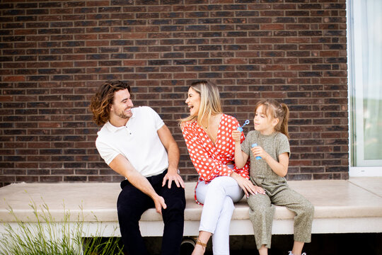 Family with a mother, father and daughter sitting outside on the steps of a front porch of a brick house - Powered by Adobe