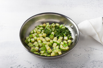 Diced fried zucchini in a frying pan with oil and chopped parsley on a light gray table. Cooking delicious vegan food