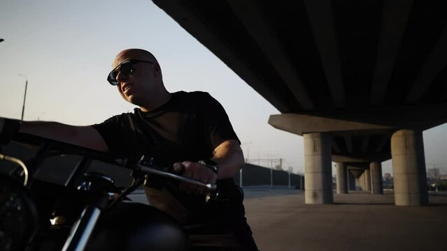 A biker sits on his favorite motorcycle with a smile. Riding a motorcycle without a helmet. Black chrome bike. A man in sunglasses. Shooting under the road bridge. Car summer parking in the evening. A