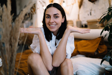 Smiling female sitting on bed and shrugging shoulders