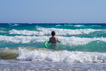Fototapeta na wymiar Girl swims with green foam swimming tube near beach of wavy mediterranean sea