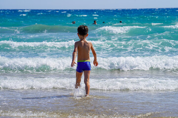 Fototapeta na wymiar Boy walking into the wavy mediterranean sea and some people are swimming blurred far away in the middle of the sea